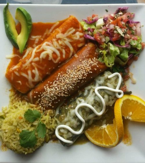 A plate of colorful Mexican food featuring enchiladas, rice, salad, and garnishes like avocado and orange slices.