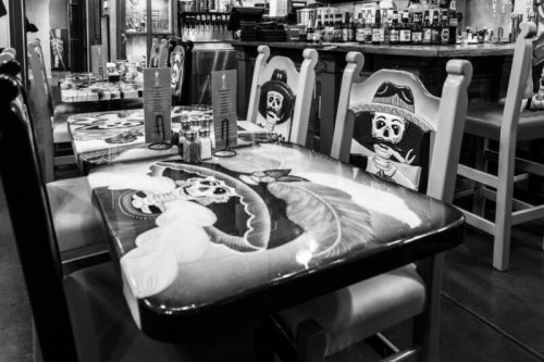 A black and white image of a restaurant table with colorful, painted chairs featuring a skeleton in a sombrero design.