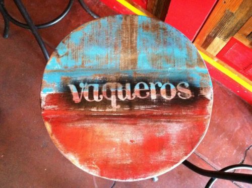 A round wooden stool with a colorful, distressed finish featuring the word "vaqueros" painted in white.