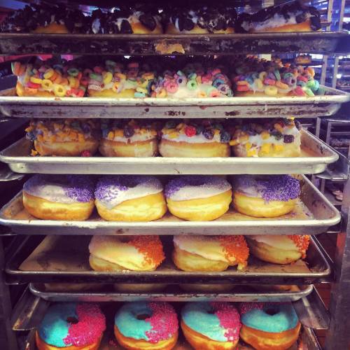 A display of colorful donuts topped with various sprinkles and cereals, stacked on metal trays.