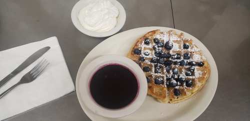 A plate with a blueberry waffle topped with powdered sugar, a bowl of syrup, and a side of whipped cream.