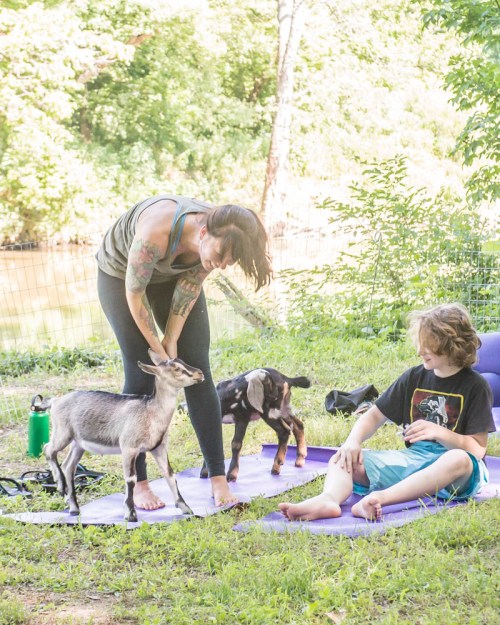 A woman interacts with a goat while a child sits nearby on a mat, surrounded by greenery and a peaceful outdoor setting.