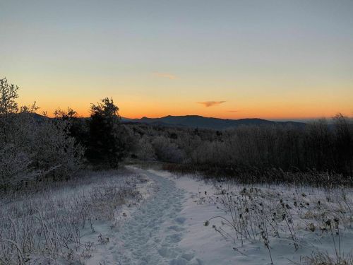 A snowy path leads through a frosty landscape at sunset, with mountains silhouetted in the background.