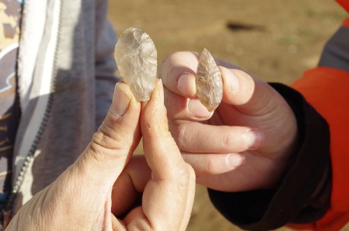Two hands hold stone tools, showcasing a larger and a smaller flint artifact against a blurred outdoor background.