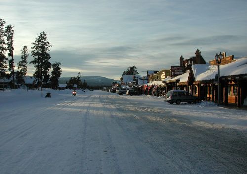 Snow-covered street in a small town, lined with shops and trees, under a cloudy sky.