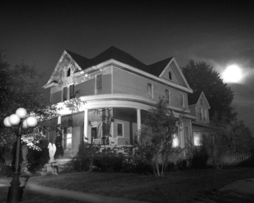 A large, old house with a wraparound porch, illuminated by moonlight, surrounded by trees in a nighttime setting.