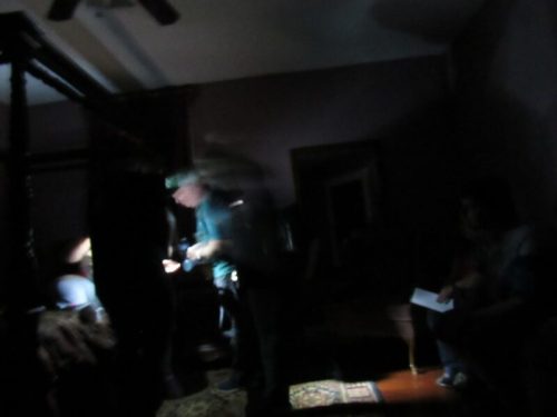 A dimly lit room with blurred figures, one person holding a light, and another seated in the background.