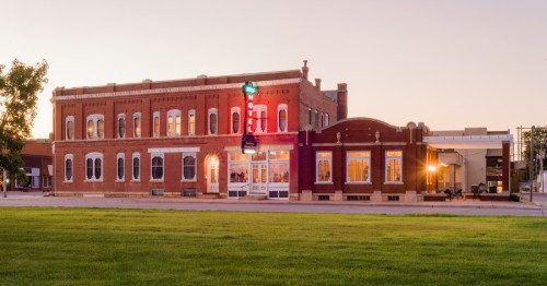 Historic brick motel building at sunset, featuring a neon sign and large windows, surrounded by green grass.