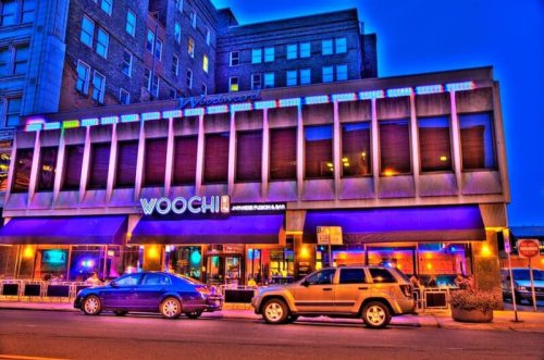 A vibrant restaurant exterior at dusk, featuring "Woochi" signage and colorful lighting, with parked cars in front.