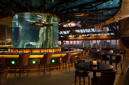 A stylish restaurant interior featuring a large aquarium, bar seating, and panoramic city views at dusk.