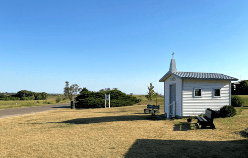 A small white chapel with a cross, surrounded by grass and trees, under a clear blue sky.