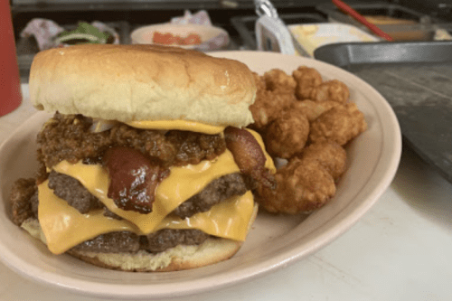 A stacked cheeseburger with bacon and chili, served with crispy fried bites on the side.