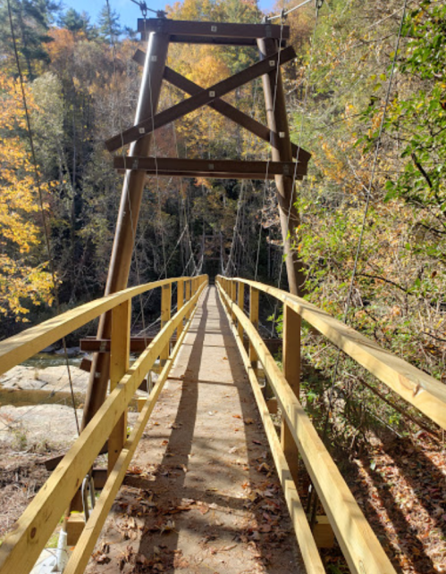 A wooden suspension bridge spans a rocky path, surrounded by autumn foliage and trees.