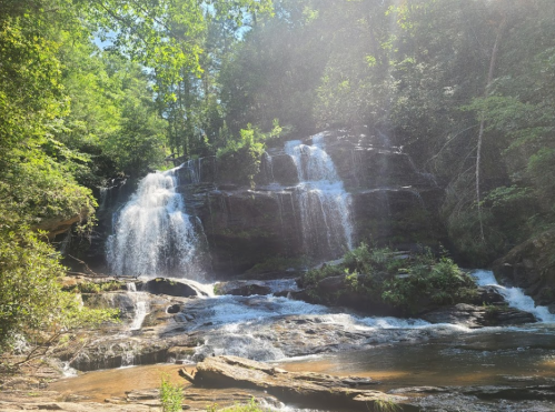 A serene waterfall cascades over rocks, surrounded by lush greenery and sunlight filtering through the trees.