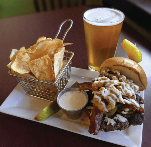 A hearty burger topped with bacon and sauce, served with crispy chips and a pint of beer on a table.