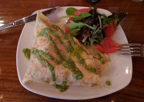 A plate with a folded crepe drizzled with green sauce, accompanied by a side salad and sliced tomatoes.