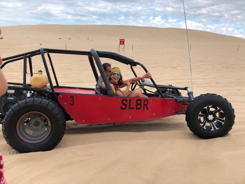 A person driving a red dune buggy on sandy terrain, with another passenger enjoying the ride.