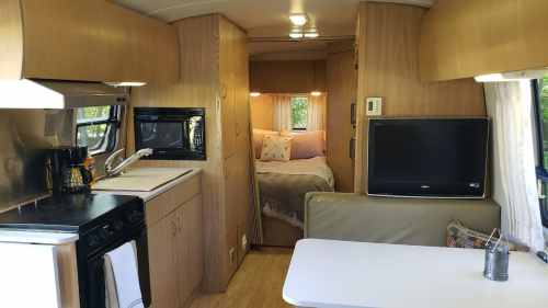Interior of a cozy camper with a kitchen, dining area, and a bedroom visible in the background. Warm wood tones throughout.