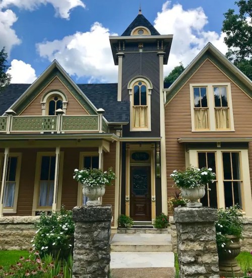 Victorian-style house with a tower, decorative trim, and a flower-filled garden under a partly cloudy sky.