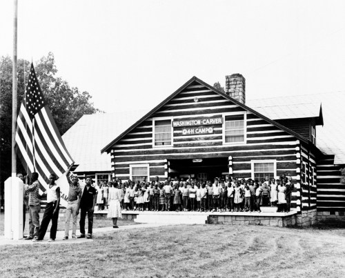 A large group of people stands in front of a log cabin building, holding an American flag, in a historic black-and-white photo.