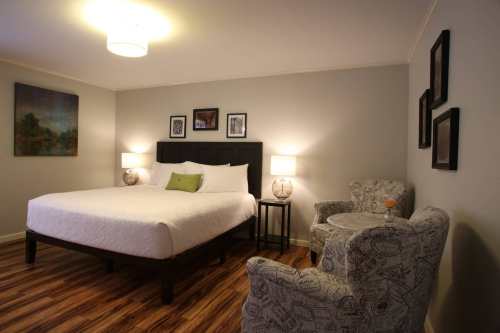 Cozy hotel room featuring a queen bed, armchair, side table, and artwork on the walls with warm lighting.
