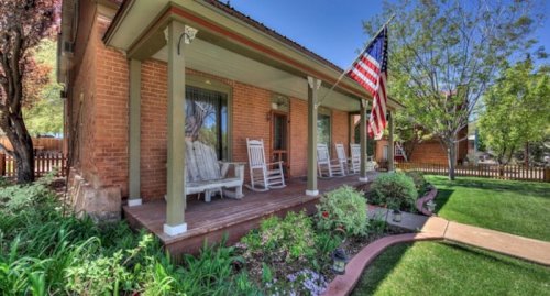 A charming brick house with a porch, rocking chairs, and an American flag, surrounded by greenery and flowers.