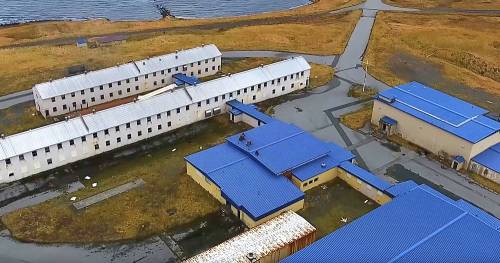 Aerial view of a complex of buildings with blue roofs, surrounded by grass and near a body of water.
