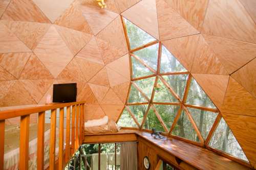 Interior of a geodesic dome with wooden paneling, large windows, and a cozy sleeping area.