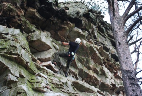A climber ascends a rocky cliff, wearing a helmet and harness, with trees in the background.