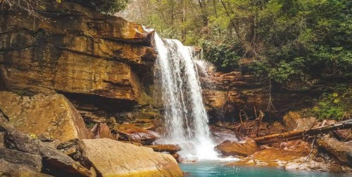 A serene waterfall cascades over rocky cliffs into a tranquil pool, surrounded by lush greenery and trees.