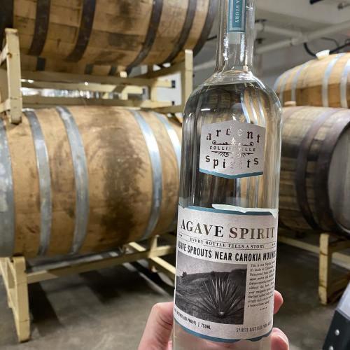 A hand holds a bottle of Agave Spirit in front of wooden barrels in a distillery setting.