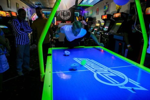 A person plays air hockey in a vibrant arcade, surrounded by spectators and colorful arcade machines.