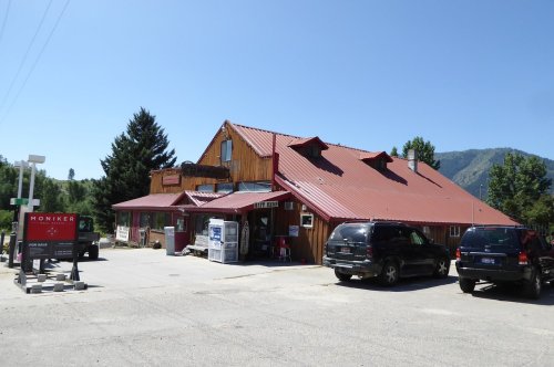 A rustic building with a red metal roof, surrounded by trees and parked cars, set against a clear blue sky.