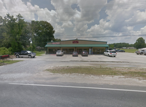 A donut shop with a green roof and sign, surrounded by parked cars and trees under a cloudy sky.