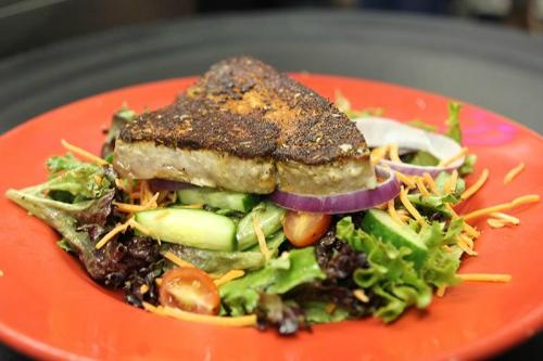 A heart-shaped piece of grilled fish on a bed of mixed greens, topped with sliced onions and cherry tomatoes.