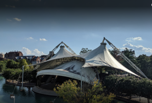 A large, white tent-like structure by a body of water, surrounded by trees and buildings under a clear blue sky.