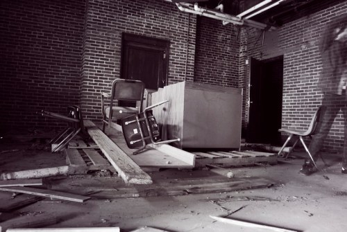 An abandoned room with overturned chairs, wooden planks, and brick walls, creating a sense of neglect and decay.