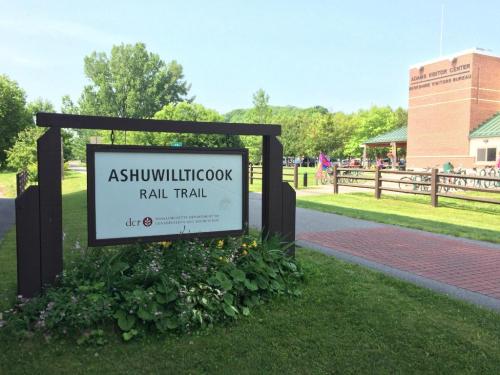 Sign for Ashuwillticook Rail Trail with a visitor center in the background, surrounded by greenery and flowers.