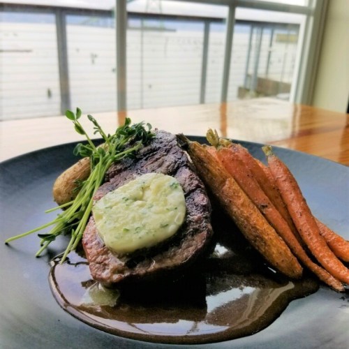 A plated steak topped with herb butter, served with roasted carrots and potatoes, against a scenic window backdrop.