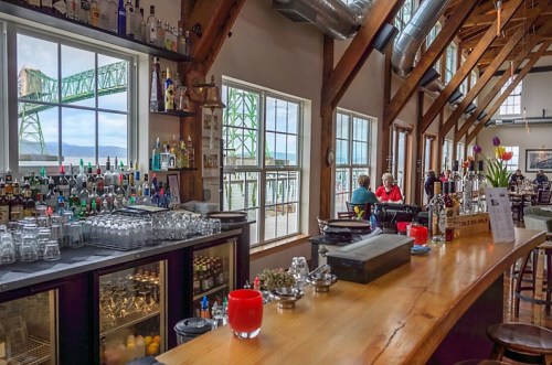 A cozy bar with a wooden counter, shelves of drinks, and large windows showing a bridge and waterfront view.