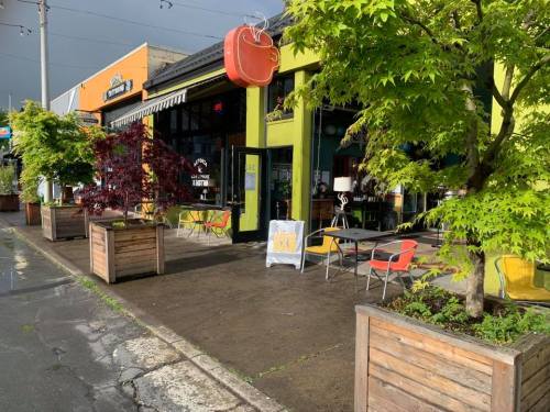 A colorful restaurant exterior with outdoor seating, surrounded by planters and trees, on a rainy day.