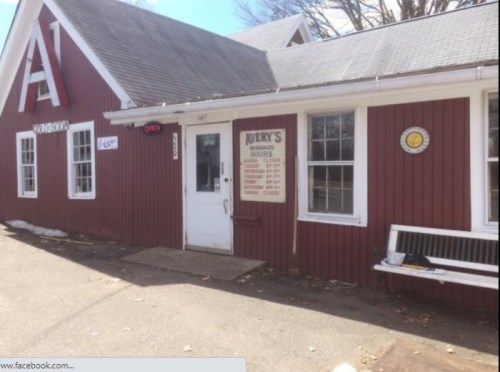 A red building with a sign reading "Nery's" and hours of operation, featuring a bench outside and a clock on the wall.