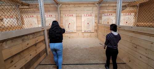 Two women stand in an indoor axe-throwing range, preparing to throw axes at wooden targets.
