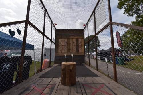 A wooden axe-throwing setup with targets, surrounded by a fence, at an outdoor event with tents in the background.