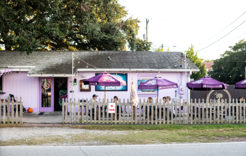 A pink restaurant with outdoor seating under purple umbrellas, surrounded by a wooden fence and trees.