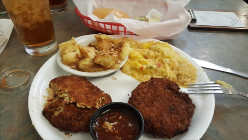 A plate of fried crab cakes with dipping sauce, served alongside cornbread, a side dish, and a drink in the background.