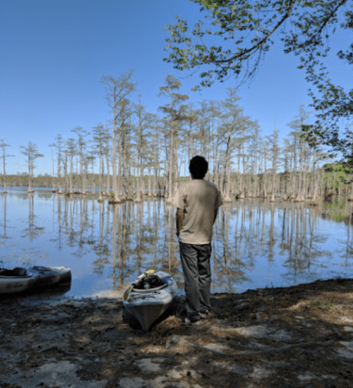 A person stands by a calm lake surrounded by trees, with a kayak nearby under a clear blue sky.