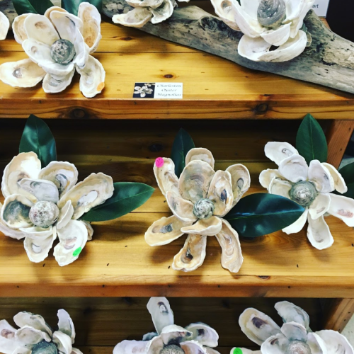 Decorative flowers made from oyster shells, displayed on wooden shelves with green leaves and labels.