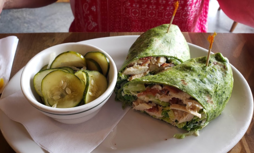 A spinach wrap filled with chicken and lettuce, served with a side of pickles on a wooden table.