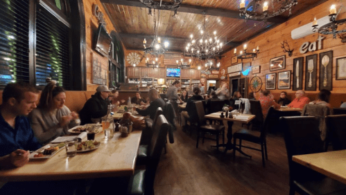 A cozy restaurant interior with wooden decor, chandeliers, and diners enjoying meals at tables.